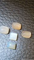 Five rectangular natural peach moonstone cabochons placed on textured black surface, showing varied shades from light peach to milky white with translucent and polished finish.
