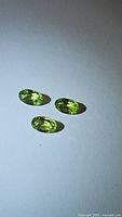 Three natural oval cut Peridot gemstones laid out on a white background with visible facets and bright apple green color.