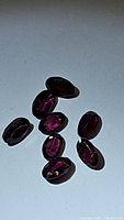 Top view of nine oval cut Rhodolite garnet gemstones showing their purple-pink color and clarity, arranged loosely on a grey surface.