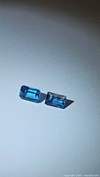 Two natural Swiss blue topaz gemstones, one emerald cut and the other baguette cut, displayed on white paper to show their color and cut details.