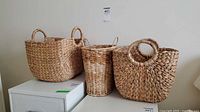 Three woven baskets displayed together on a white surface against a wall, showing relative size and handle styles.
