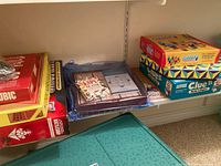 Photo showing stacked board games including Qubic, Checkers, Electronic Battleship, and smaller boxed games like Sudoku and Sushi Go Fish card game.