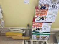 Image showing two boxed sets of FoxRun butter warmers stacked against the wall alongside ceramic corn holders, fish-shaped plates, and glass dispenser bottle on kitchen counter.