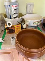 Photo showing the fondue set components: a large brown ceramic bowl, a smaller cream-colored ceramic bowl with handle, two tubes of Starfrit liquid fondue concentrate, and a box for fondue chocolate, all placed on kitchen counter