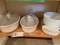 Shelf with various white CorningWare ceramic casserole dishes, some with glass lids, in different sizes and shapes, all clean with no visible chips or cracks.