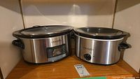 Two Crock-Pot slow cookers side by side on a shelf, one digital, one manual dial, both stainless steel with glass lids.