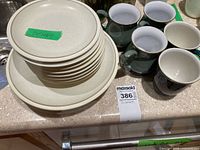 Set of stacked beige/light cream colored stoneware plates and matching mugs on countertop labeled Denby.