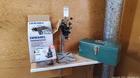 Dremel shaper/router table box, metal toolbox, and metal base accessory pictured on white shelf against plywood wall.