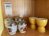 Photo showing four Royal Worcester ceramic egg coddlers with metal lids and two yellow ceramic egg cups on wooden shelf.