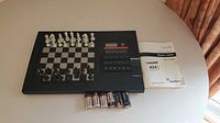 Overall view of the RadioShack Master 2200X electronic chess computer with chess pieces set on the board, instruction manual, box, and batteries on a round table.