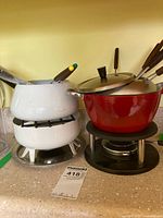 Two fondue sets side by side, red and white pots with burners underneath and forks inside.