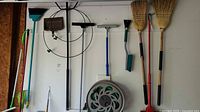 Photo of various garden tools hanging and leaning against a wall including brooms, rakes, brushes, and coiled garden hose reel.