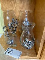 Full view of 4 glass vases with metal floral and pewter accents arranged together on wooden cabinet shelf.