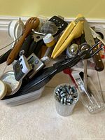 Photo of assorted kitchen utensils including wooden spoon, plastic tongs, yellow citrus juicer, metal thermometer, and digital timers in a clear plastic container