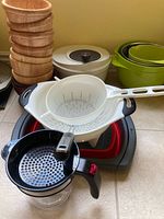 Photo showing stacked wooden bowls, white plastic colander, nested green bowls, and a black handled kitchen tool with clear container and crank handle.
