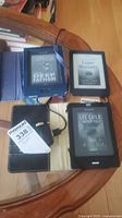 Three Kobo eReaders placed on a table, showing ebook covers on their screens. Protective covers and a charger visible.