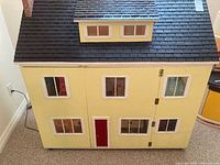 Exterior front view of the large wooden dollhouse painted yellow with black shingled roof, multiple windows, and red front door.