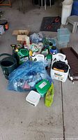 Overall image showing collection of lawn and garden chemicals and tools on concrete floor including bottles, containers, sprayers, and various accessories.
