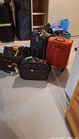 Several pieces of assorted luggage grouped on a basement floor, including black, navy, and orange soft-sided bags with wheels and handles visible.