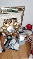 Overview of assorted mirrors, including rectangular wall mirrors with white and silver plastic frames, a gold sun-shaped mirror, tabletop mirrors, handheld mirrors and magnifying glasses on wooden floor.