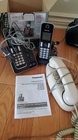 Panasonic KX-TG393C cordless phone with base and handset, Aastra Sabrina white corded phone, Panasonic instruction manual and cords on floor.