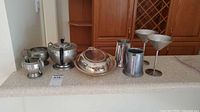 Full view of all pewter barware and tea set items arranged on counter including tray, teapot, sugar bowl, creamer, two stemmed glasses, and two tankards.