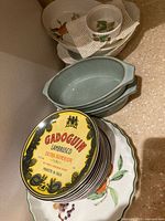 Overview of ceramic dishes showing Evesham Vale Royal Worcester fruit motif plates, sky-blue Schumann Arzburg casseroles, and decorative wine label plates stacked