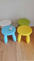Four plastic child stools in white, light blue, green and yellow arranged on wooden floor against white wall.