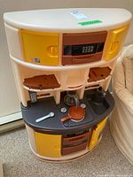 Front view of plastic play kitchen showing microwave, cabinets, stovetop with controls, oven, and sink with faucet; includes coffee pot, frying pan, spoon, and phone remote replica.