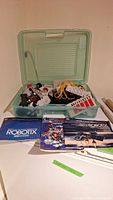 Open green plastic storage case showing various Robotix modular building parts, wires, wheels, and manuals in front of the case.