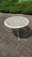 White wrought iron patio side table shown outdoors on brick patio with detailed filigree design on the round top and three curved legs.