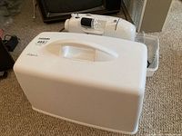 Closed white hard cover sewing machine case with handle on top, placed on carpeted floor next to a TV stand and other items.
