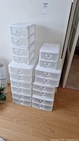 Two stacks of white plastic stackable drawer organizers with transparent drawers on wood floor against white wall. Various drawer sizes.