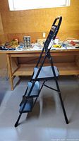 Side profile of the black metal step ladder with three silver steps showing its folding mechanism and rubber feet.