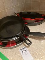 Stack of pans including cast iron skillet, nonstick frying pans, grill pan with ridges, and sauté pan with lid photographed on kitchen countertop.