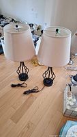 Pair of table lamps with black metal bases and white fabric shades placed on wood floor. Both lamps have cords and bulbs attached.
