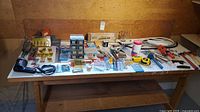 Wide shot of entire tools and hardware lot on wooden bench, showing drill, soldering iron, bits, saws, files, wrench, fasteners, hose.