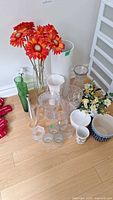 Lot of vases, planters and artificial flowers arranged on floor showing mix of glass, ceramic, and metal items with two bunches of artificial flowers (red-orange and white-yellow).