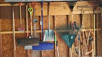 Wall-mounted assortment of long-handled yard and garden tools including shovels, rakes, snow shovels, hoes, and mattocks.