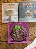 Three baking cookbooks laid flat on wooden surface: Baking with Less Sugar, Baked Occasions, and Dorie's Cookies