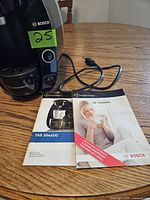 Front and side view of Bosch Tassimo TAS 20xxUC coffee maker on wood table with power cord and two manuals.