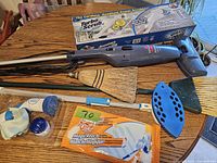 Photo showing Bissell cordless handheld power scrubber with box, broom, dustpan, mop head and handle, and smaller cleaning scrubbers and brushes.