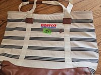 Front view of cooler bag showing Costco Wholesale logo, striped exterior pattern, cream-colored dual handles, and brown leather accent patches.