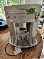 Front view of silver DeLonghi Magnifica espresso machine on wooden table, showing control knobs, indicator lights, brew group and steam wand