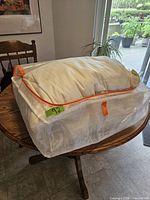 Queen size duvet inside a semi-transparent white mesh protective bag with orange zipper and handle, placed on a wooden table near a window.