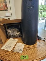 Tall black Germ Guardian air purifier unit standing next to the opened box containing replacement filter sealed in plastic and the instruction manual on a wooden table.