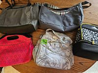 Set of five various lunch bags including gray Bench tote, gray marble fabric backpack style, red neoprene with geometric pattern, blue crossbody bag, and black Alite cooler bag.
