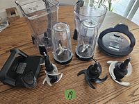 Ninja blender replacement parts laid out on a wooden table including clear blending jars, blending cups with blades, and black plastic lids.