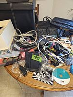 Photo shows a black wired keyboard partially visible, a pile of assorted computer cables and adapters, a small black pouch, and some labels and papers on a wooden surface.