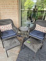 Two dark brown wicker patio chairs with navy blue seat cushions and patterned lumbar pillows, two matching ottomans, and a small round side table between the chairs outside on patio tiles.